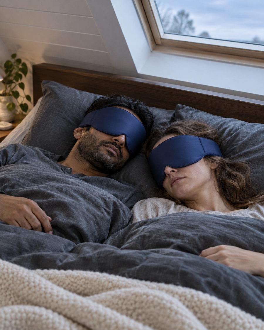 Casal dormindo lado a lado na cama, cada um com uma máscara de seda azul-marinho envolvente. Imagem ilustrativa
