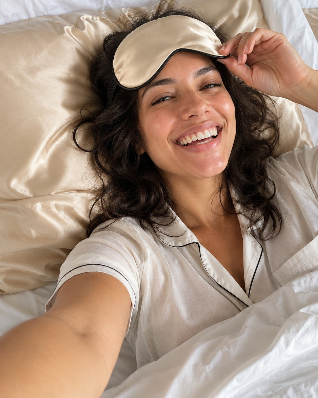Mulher sorrindo com a máscara de seda champanhe na testa, deitada em lençol branco. Imagem ilustrativa