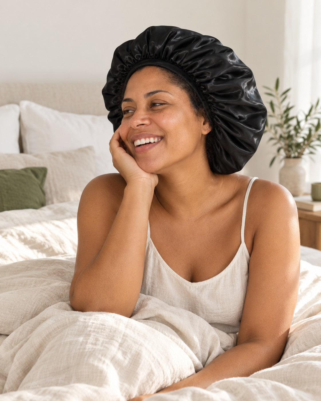 Mulher sorrindo na cama de manhã, com a touca de cetim preta na cabeça. Imagem ilustrativa.